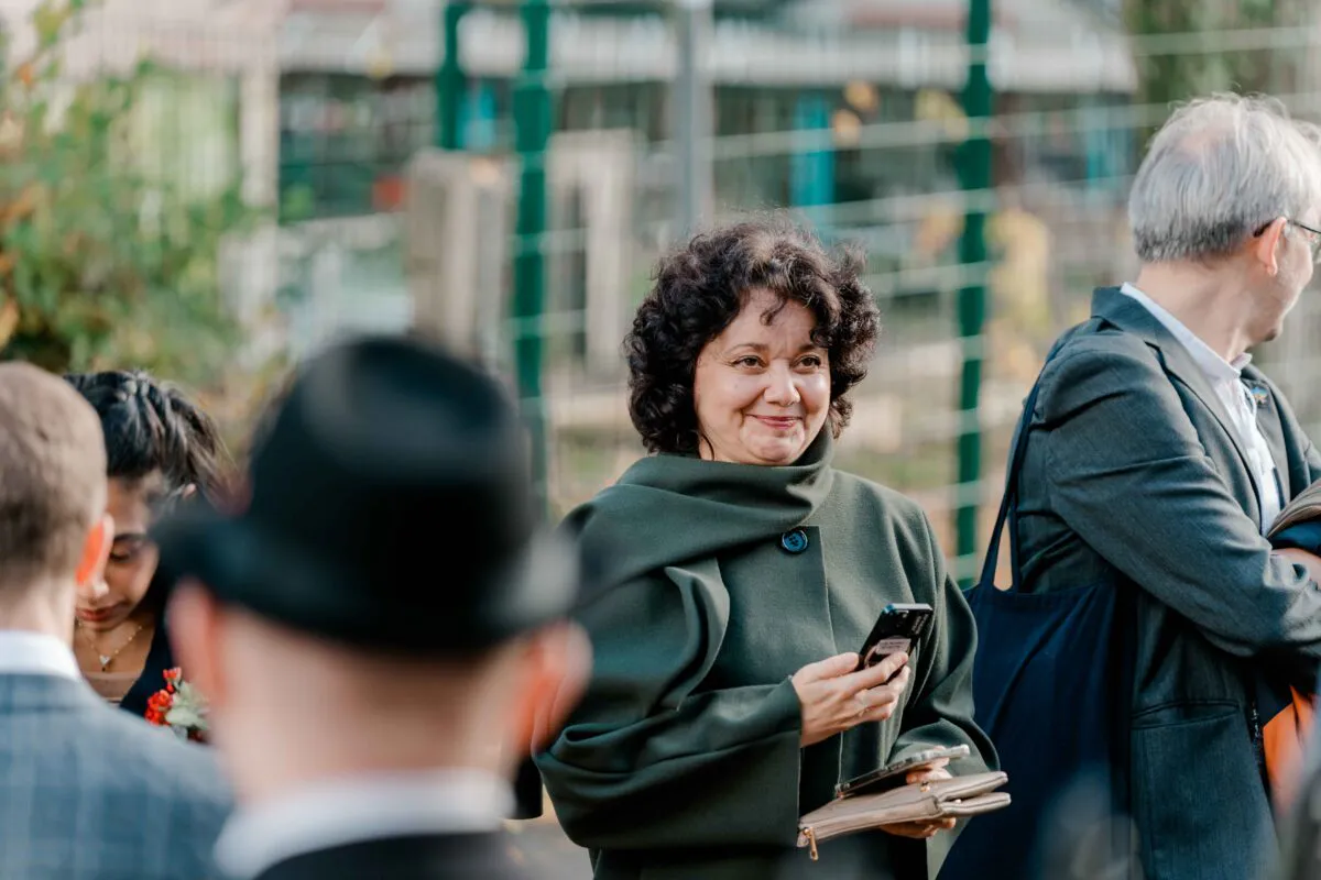 A smiling guest with dark curly hair wearing a dark green coat holds a mobile phone while mingling at an outdoor wedding celebration.