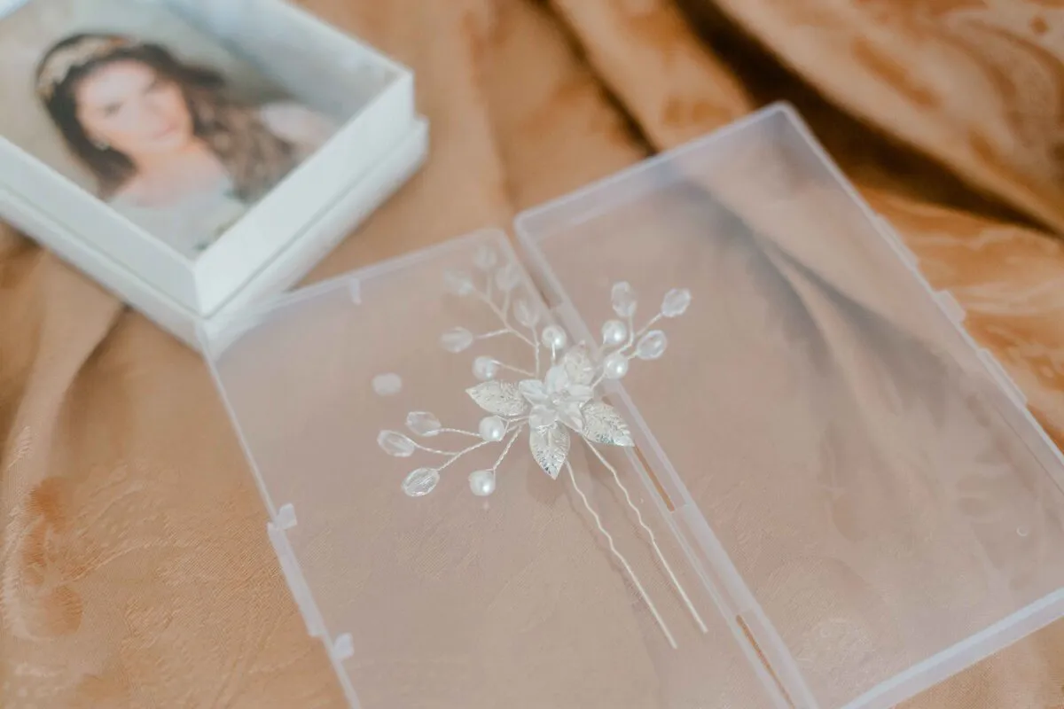 A delicate silver bridal hair pin featuring pearls and leaf details rests inside a clear plastic box next to an open white box containing a framed photo of a bride.