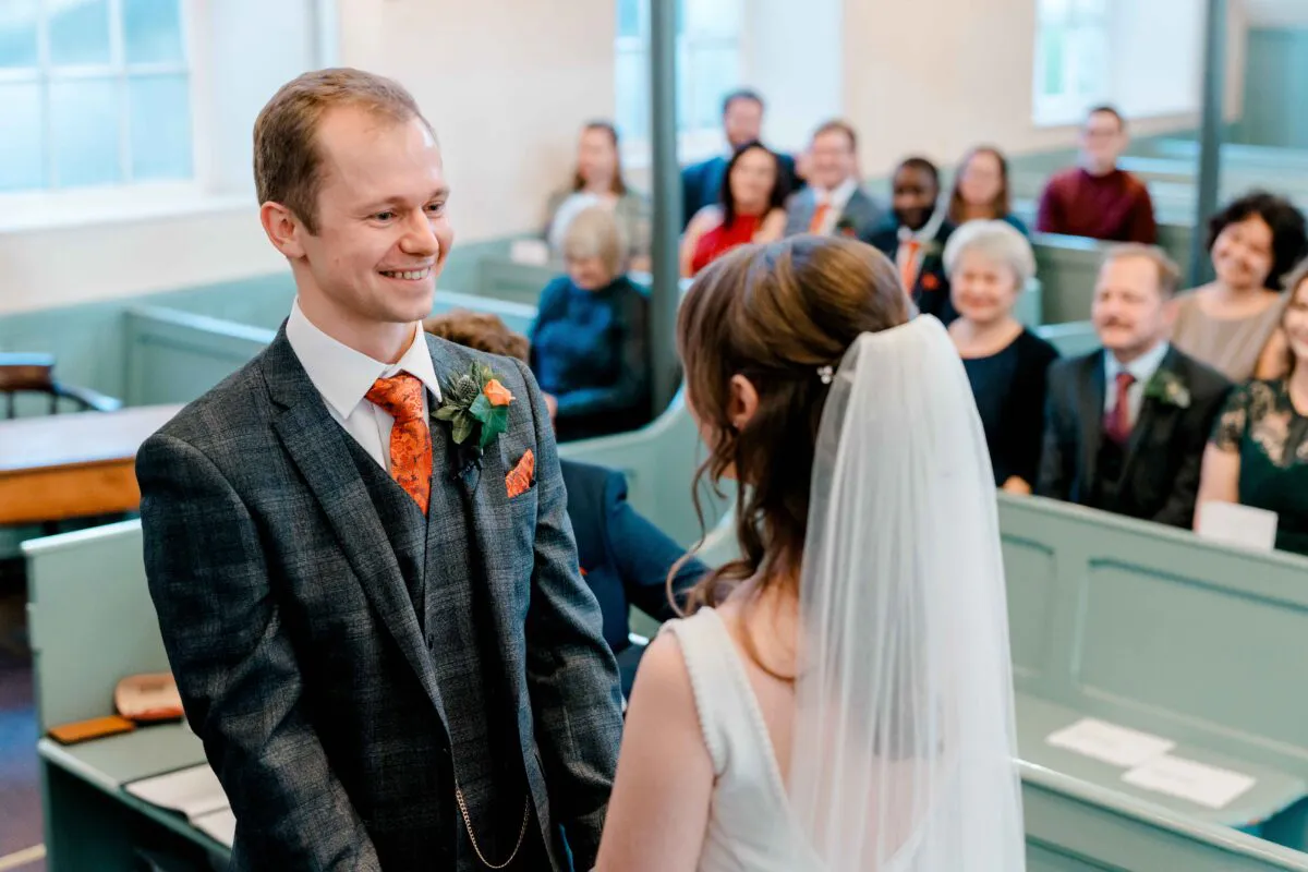 A smiling groom looks lovingly at his bride during their indoor chapel wedding ceremony while seated guests watch from the pews behind them.