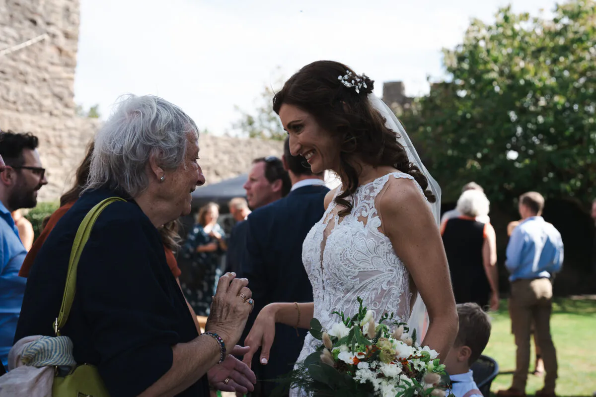 A smiling bride in an illusion neckline lace gown holding a bouquet chats with an older guest during an outdoor wedding reception.