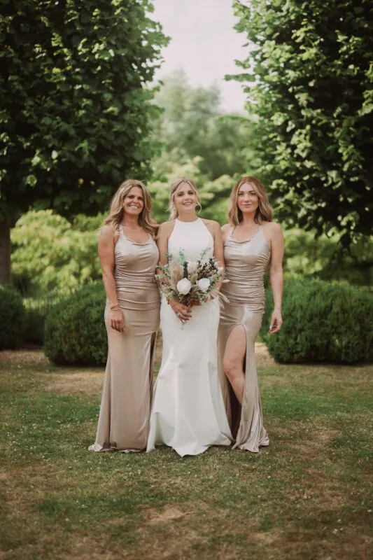 A smiling bride holding a dried flower bouquet stands flanked by two bridesmaids wearing matching champagne satin dresses with thigh splits in a lush garden setting.