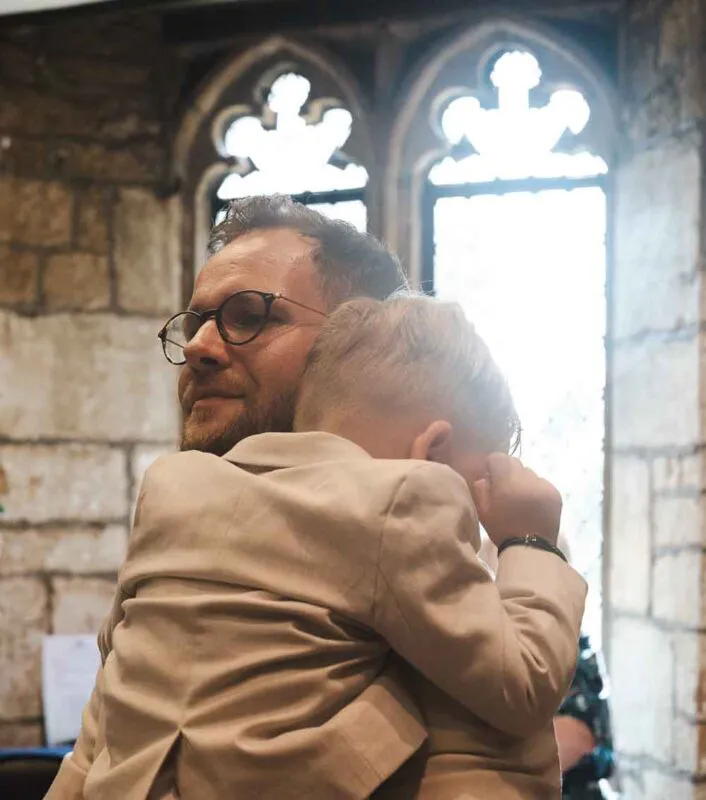 A man wearing glasses and a beige suit holds a young page boy in his arms inside a historic stone church with stained glass windows.