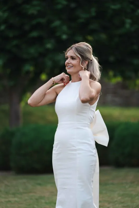 A smiling bride wearing a chic halter neck column dress with an oversized bow detail stands outdoors amidst lush green scenery.