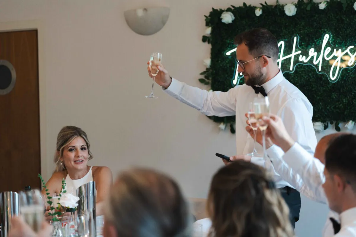 A groom in a white shirt and bow tie raises a glass of champagne during wedding speeches while the bride watches and smiles from the top table.