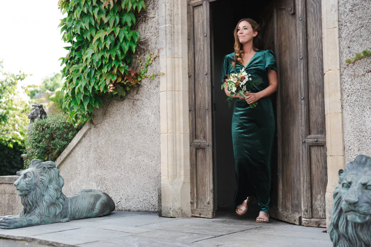 A smiling bride wearing an emerald green velvet dress and holding a small bouquet stands in a rustic wooden doorway flanked by stone lion statues.