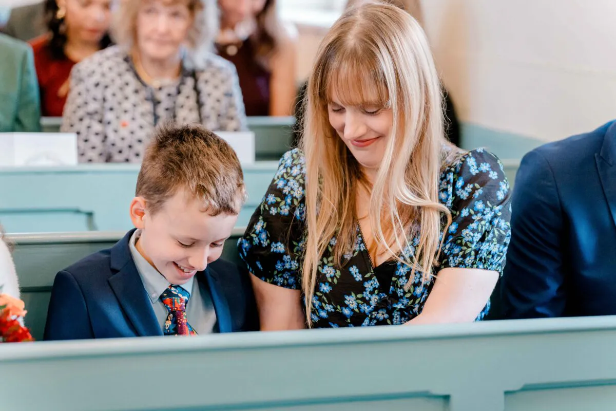 A young boy and a woman share a joyful, smiling moment while seated in wooden pews during a wedding ceremony.