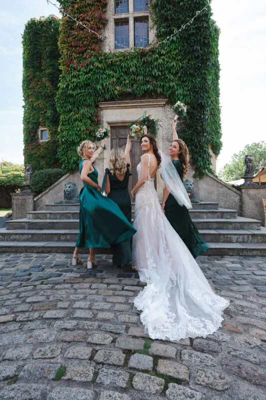 The bride and her bridesmaids pose joyfully on cobbled stone steps in front of a majestic, ivy-covered stone castle tower.