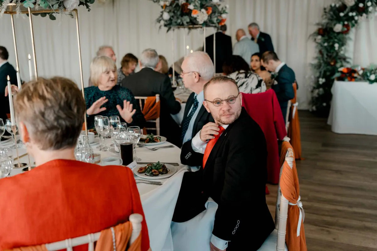 Guests enjoy a lively wedding breakfast seated at round tables decorated with burnt orange accents and tall floral centerpieces.