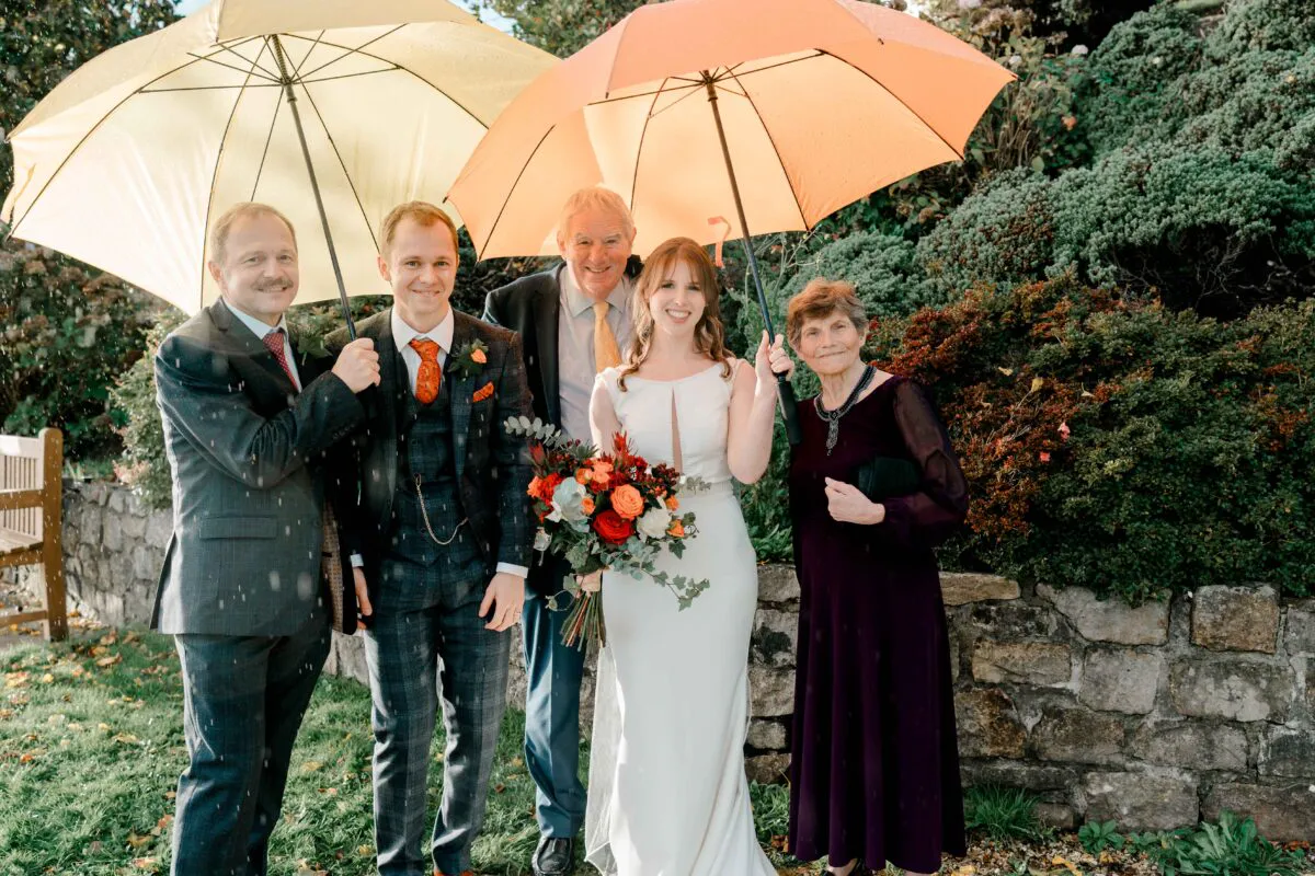 A happy wedding party consisting of a bride, groom, and family members pose outdoors beneath colourful yellow and orange umbrellas during a rainy autumn day.