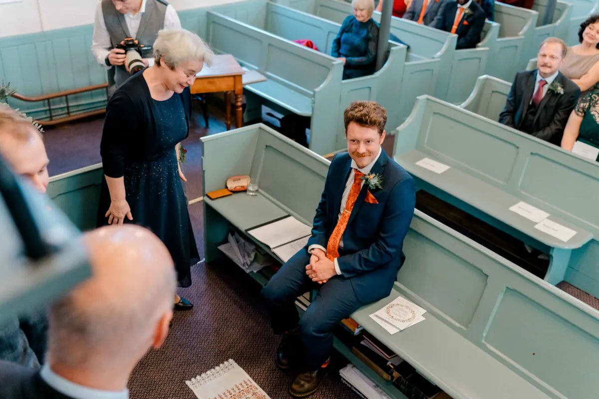 A groom in a navy suit and patterned orange tie sits in a wooden chapel pew while a woman in a dark dress looks down at him.