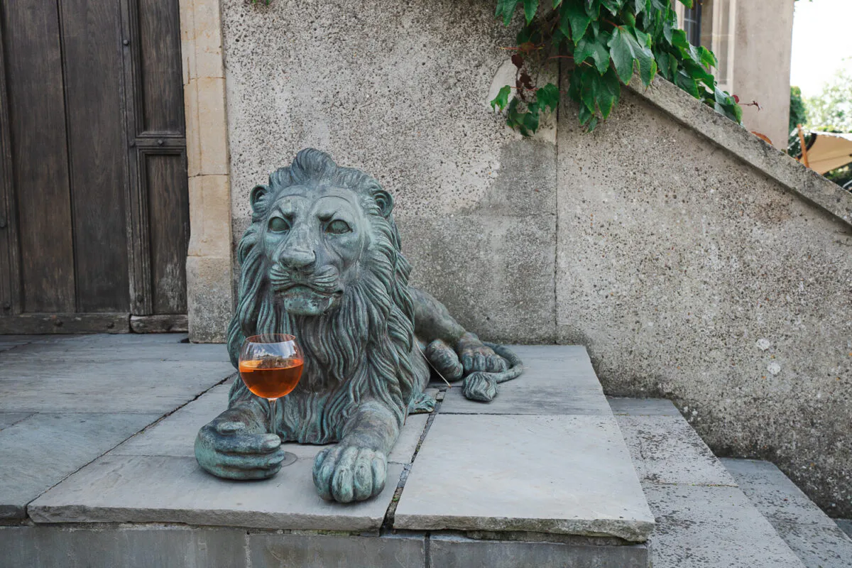 An outdoor drinks reception detail shot showing a glass of orange cocktail resting in the paw of a weathered bronze lion statue outside a stone building.