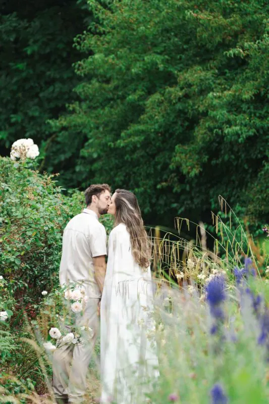 A newlywed couple shares a tender kiss while walking hand in hand through a lush green English country garden surrounded by wildflowers.