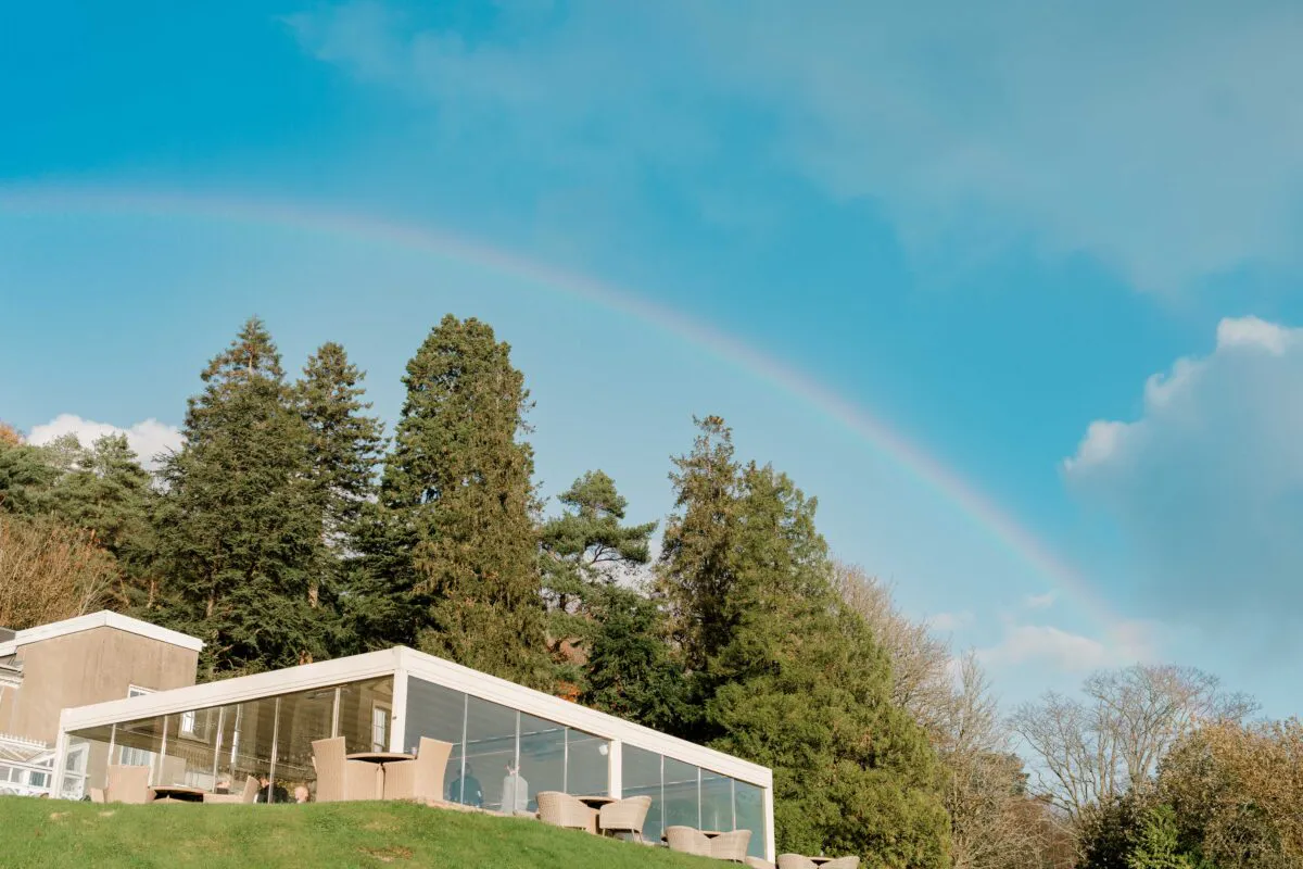 A vibrant rainbow stretches across a clear blue sky above a modern glasshouse wedding venue surrounded by tall evergreen trees and manicured lawns.