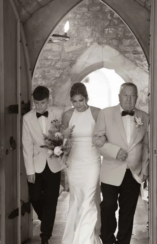A bride in a sleek halter-neck gown walks through a stone archway holding a bouquet, escorted by family members in smart suits and bow ties.