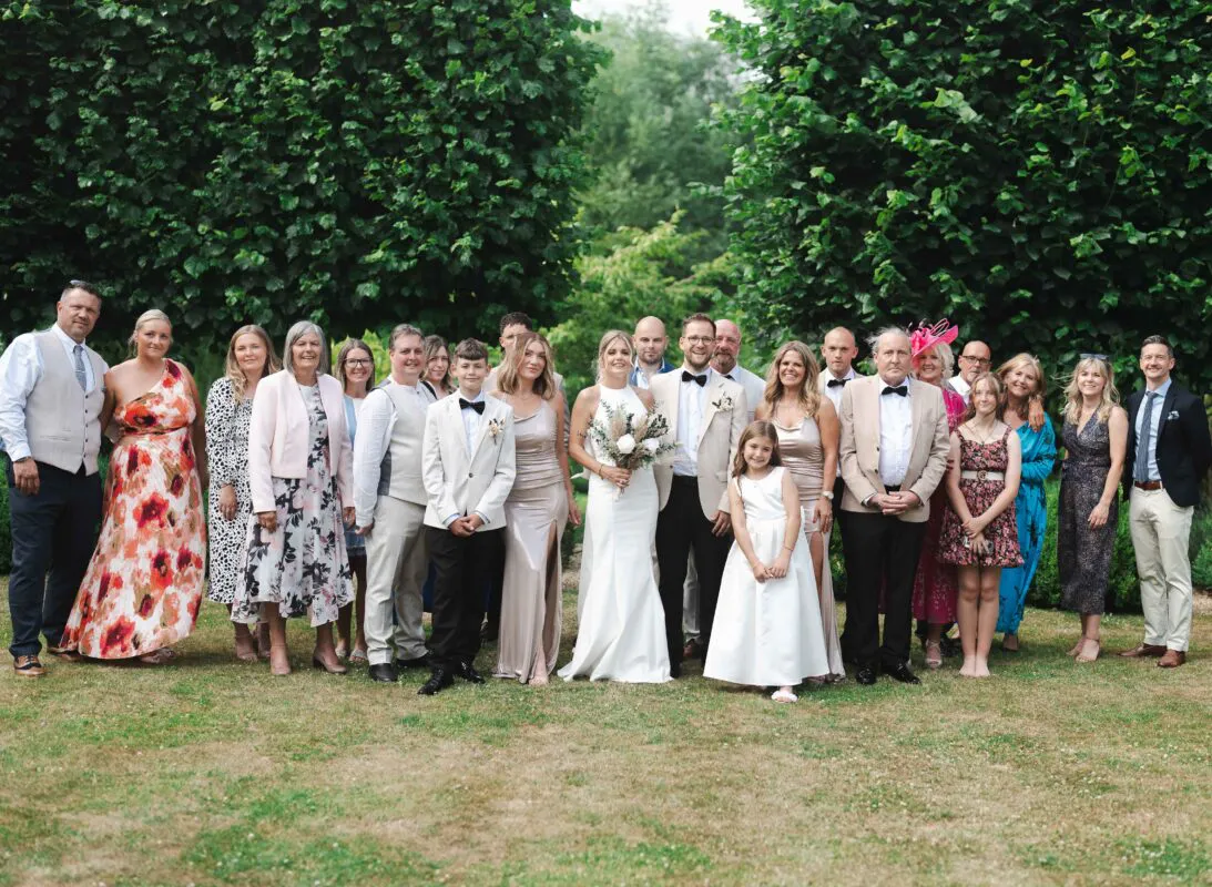 A large wedding party and family group pose together outdoors on a manicured lawn against a lush green garden backdrop.