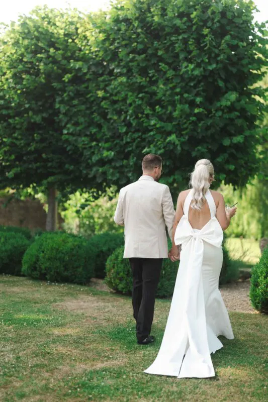 Newlyweds walk hand in hand through a lush garden, the bride wearing an elegant backless gown with an oversized bow and a high ponytail.