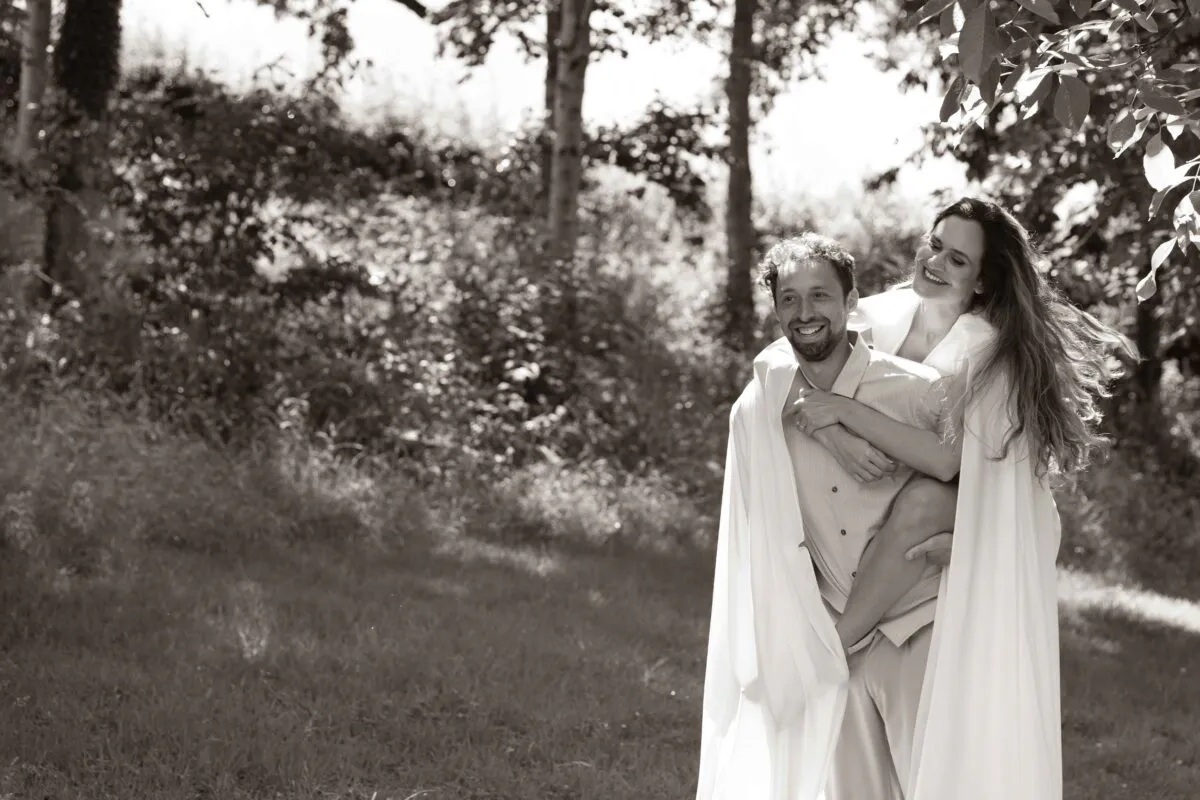 A newlywed couple shares a joyful piggyback ride amidst a sunlit woodland backdrop during their outdoor wedding portraits.