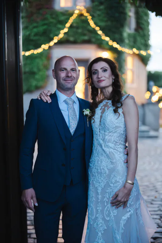 A newlywed couple poses together in a cobbled courtyard at twilight, with the groom wearing a navy three-piece suit and the bride in a detailed lace gown beneath string lights.