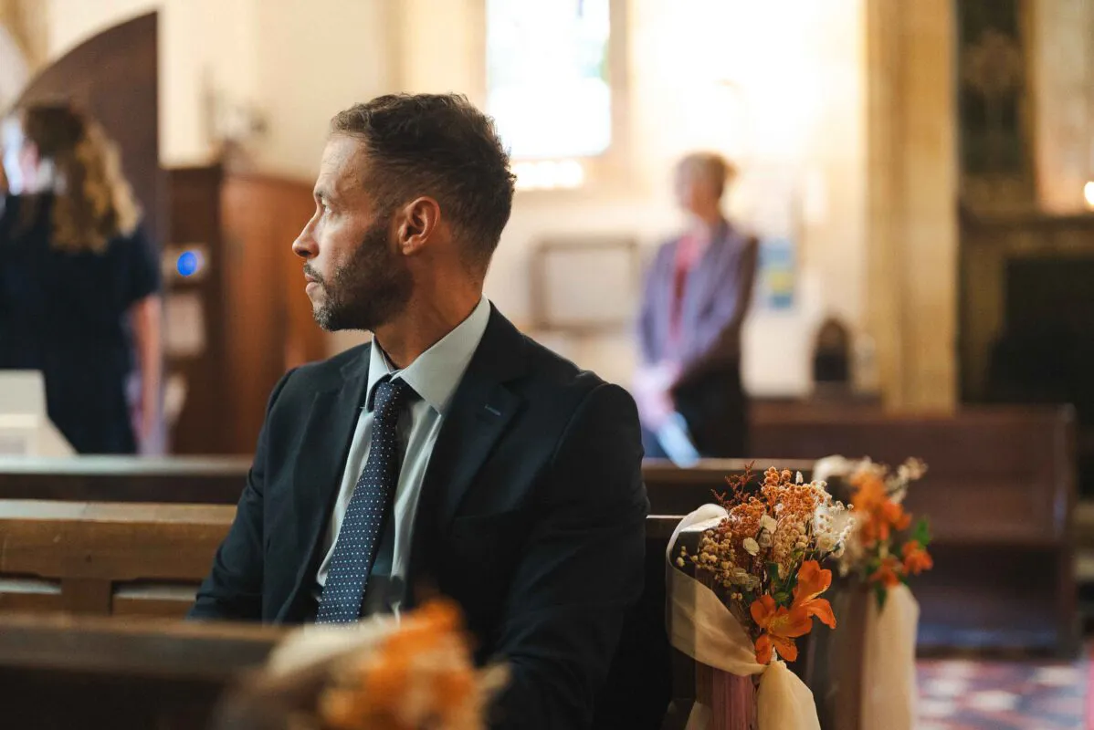 A groom in a dark suit and patterned tie sits in a church pew decorated with floral pew ends, waiting attentively during the ceremony.