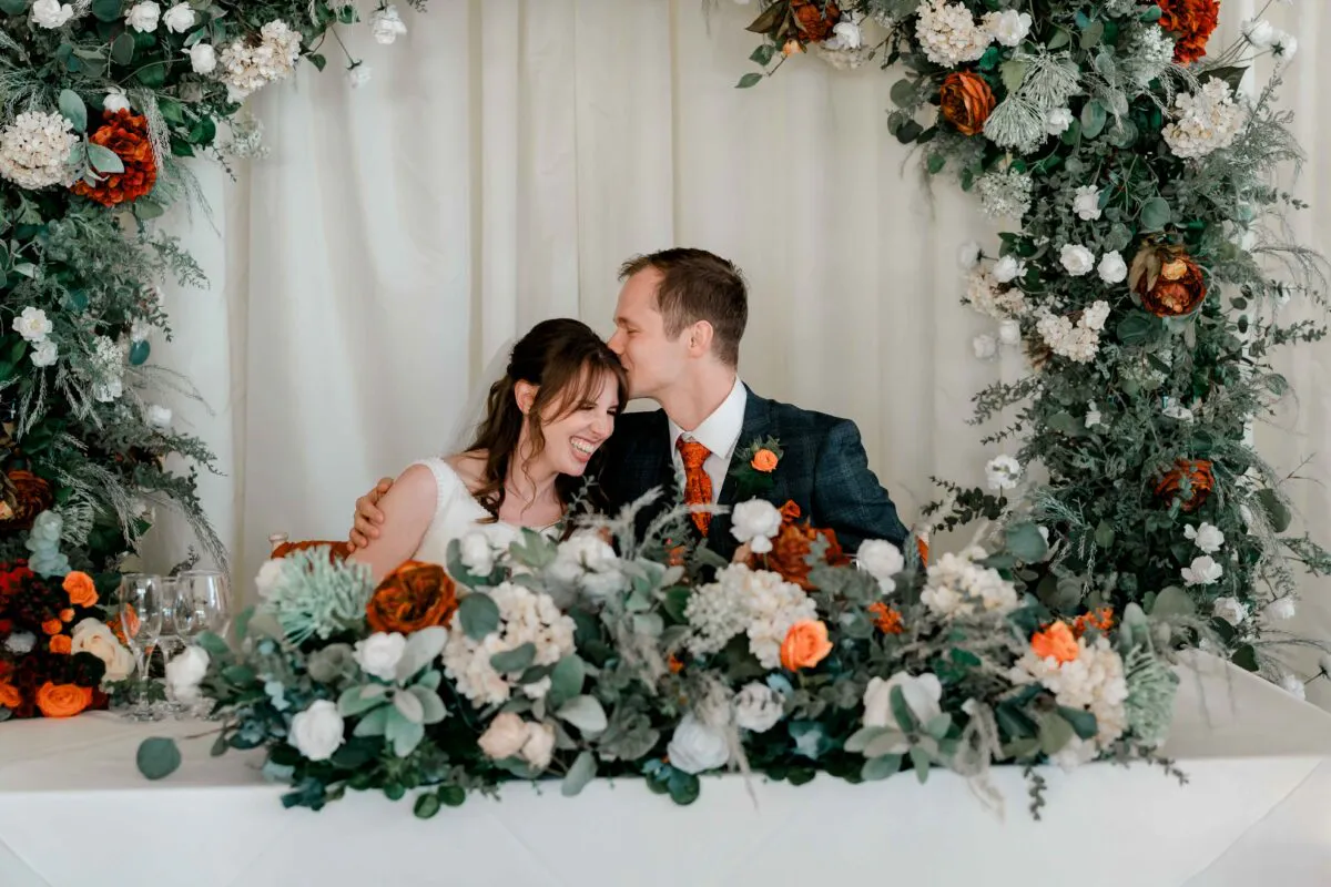 A joyful bride and groom share a tender forehead kiss while seated at a decorated top table framed by an abundant floral arch featuring rich burnt orange and white blooms.