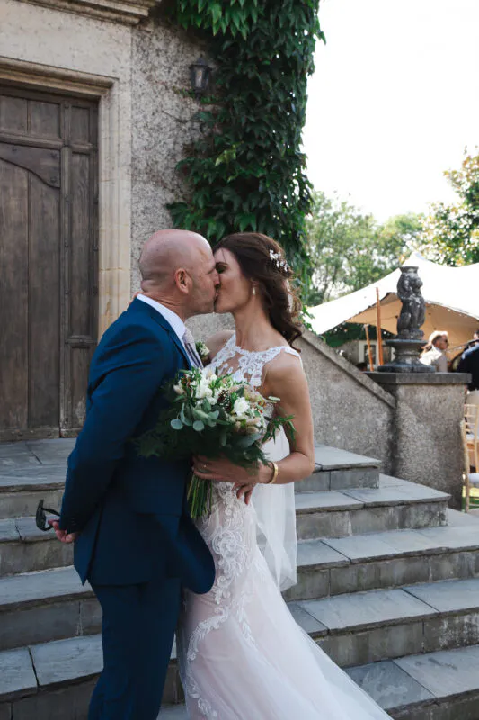 A newlywed couple shares a tender kiss on stone steps outside a historic building draped in lush ivy, with the bride holding a hand-tied bouquet.