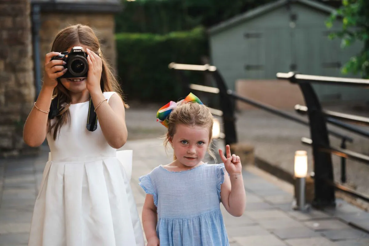 Two young wedding guests enjoy the reception, with one child playfully taking a photograph with a camera while the other flashes a peace sign.