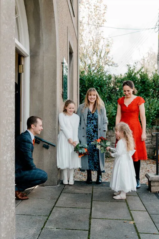 A groom in a checked suit crouches on a stone patio outside a building, smiling at a young flower girl holding a small bouquet while guests look on.