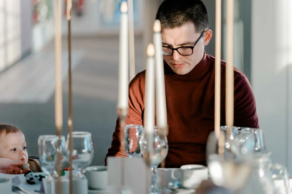 A man wearing a burgundy turtleneck and glasses sits at a wedding reception table adorned with tall taper candles and glassware, while a baby looks on from the side.