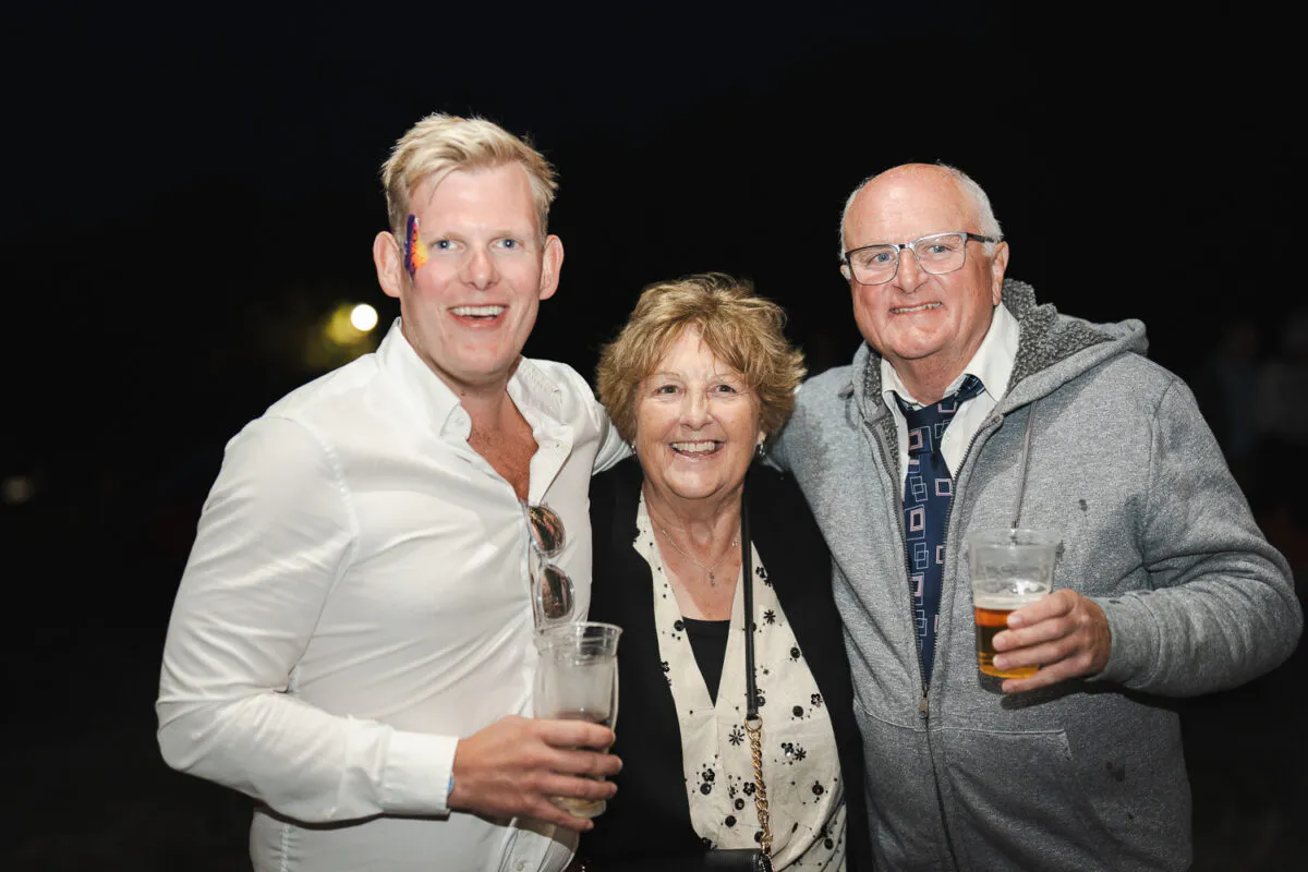 Three wedding guests laughing together at night during the evening reception, holding plastic cups of drinks.
