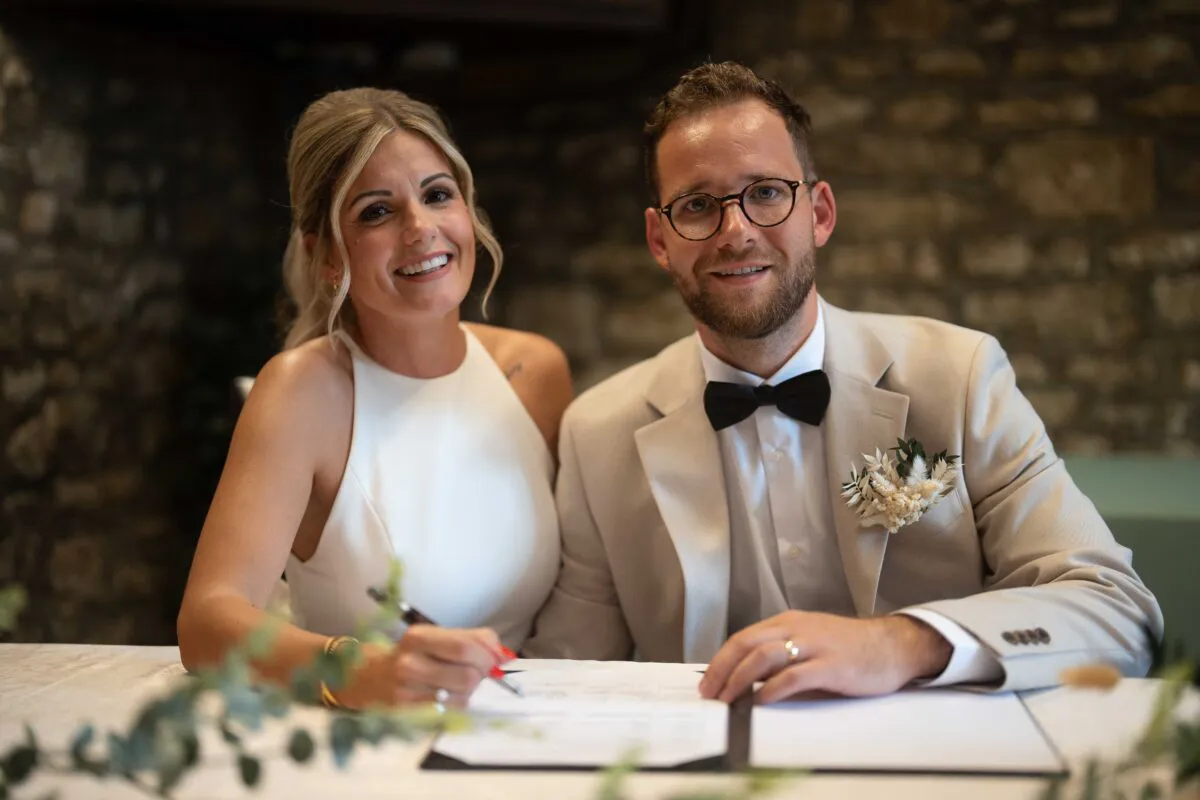 A smiling bride and groom sitting together at a table inside a stone barn while signing their wedding register.