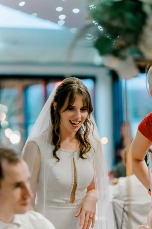 A smiling bride with half-up wavy hair and a pearl-trimmed dress laughs during a wedding reception.