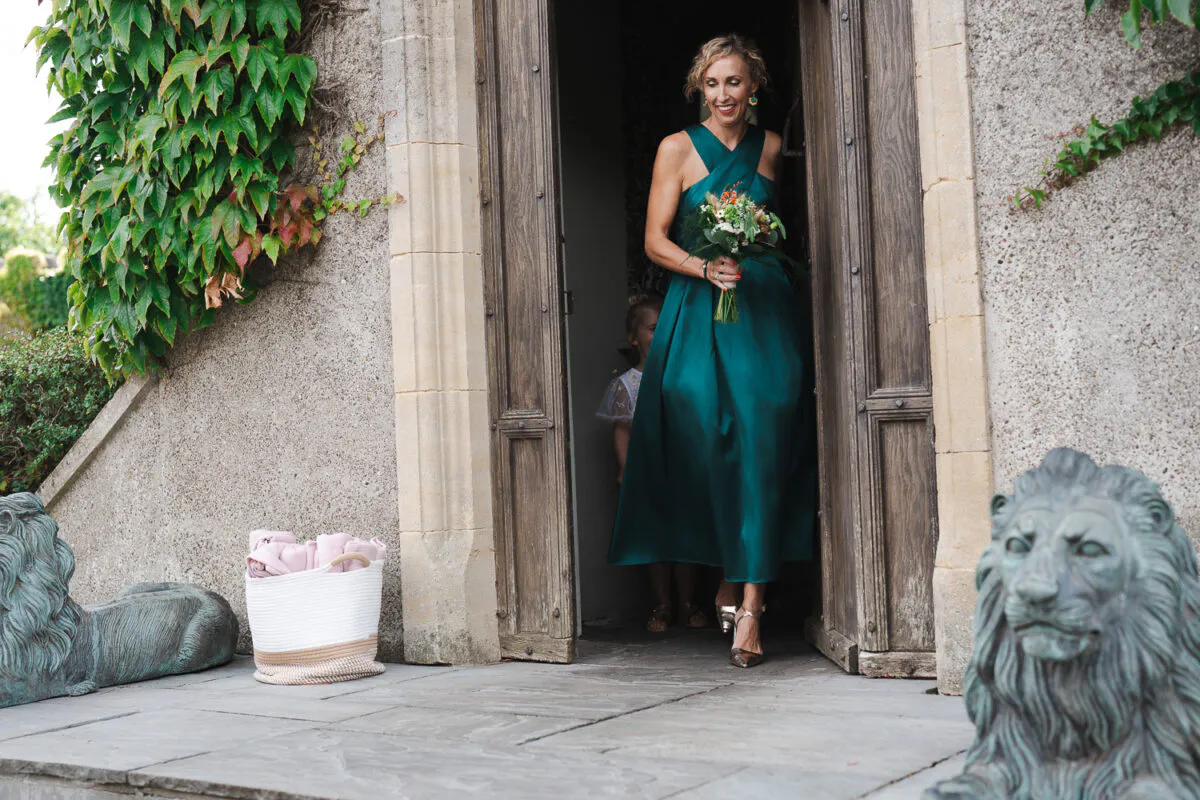 A mature bride wearing an emerald green satin gown and holding a bouquet smiles as she walks through a stone doorway flanked by lion statues.
