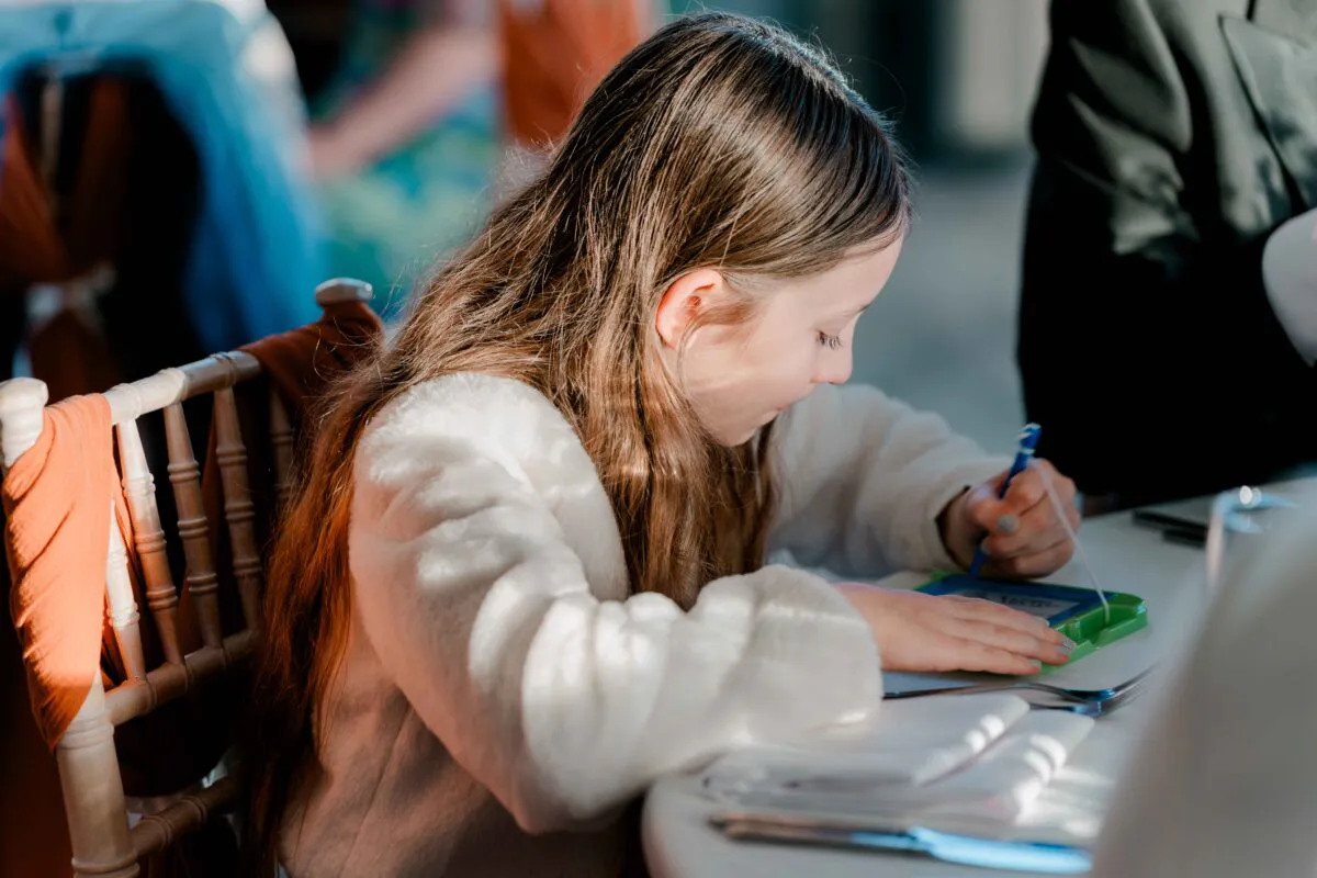 A young girl with long brown hair sits attentively at a wedding reception table, drawing with a blue pen.