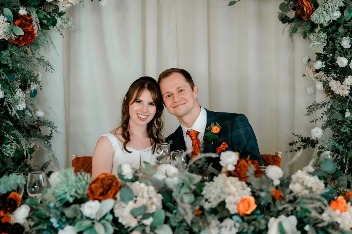A smiling bride and groom sit together at the top table, framed by an autumnal floral arch with burnt orange and white blooms.