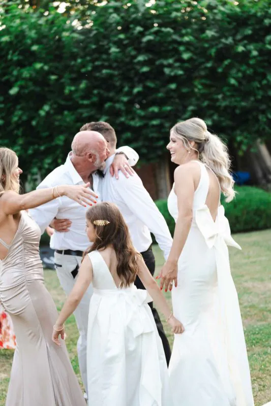 A joyous bride in a halterneck gown with an oversized bow stands outdoors alongside a flower girl and celebrating family members amidst lush green trees.