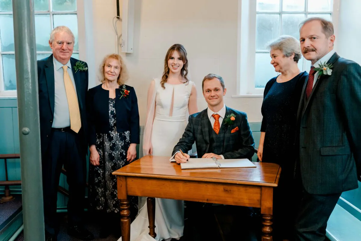 The groom sits at a wooden table signing the register while the smiling bride and family members stand grouped around them in a bright room.