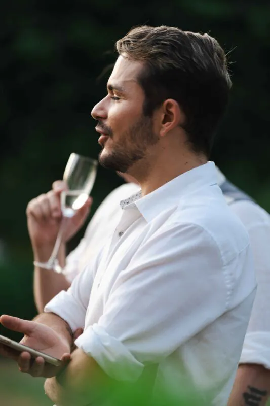 A guest in a crisp white shirt holds a champagne flute while talking and mingling during an outdoor wedding drinks reception.