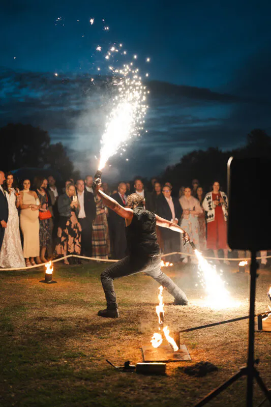 A fire performer puts on an energetic show with sparks flying into the night air while wedding guests watch from the sidelines outdoors.
