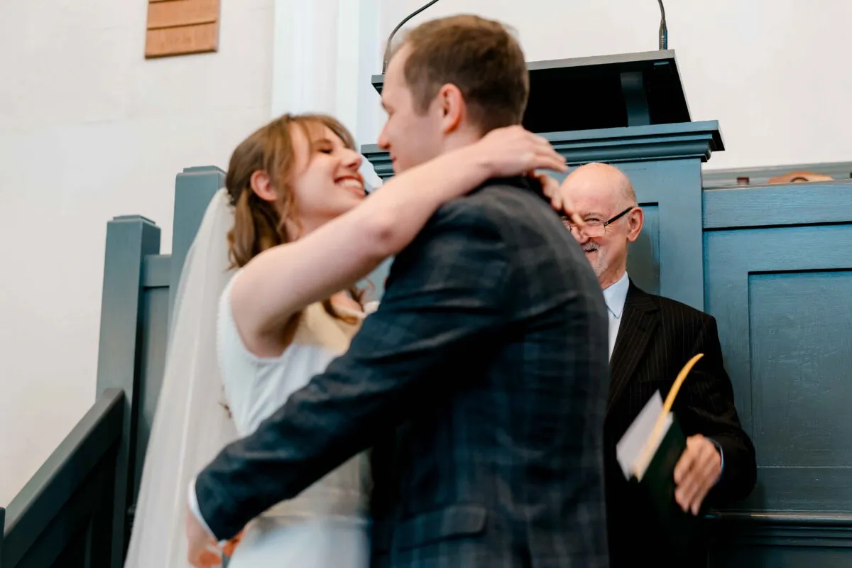 A newlywed couple embrace and laugh joyfully at the altar during their church wedding ceremony while an officiant looks on smiling.