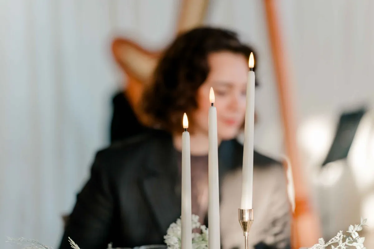 Three slender lit taper candles stand in the foreground with a softly blurred harpist playing in the background.