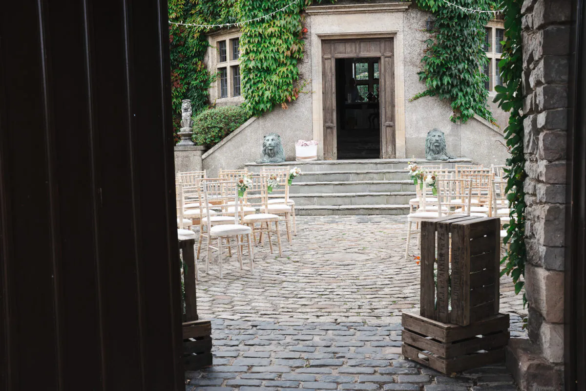 An outdoor wedding ceremony setup in a historic stone courtyard featuring a cobbled floor, rows of chiavari chairs adorned with floral pew ends, and ivy climbing the building's facade.