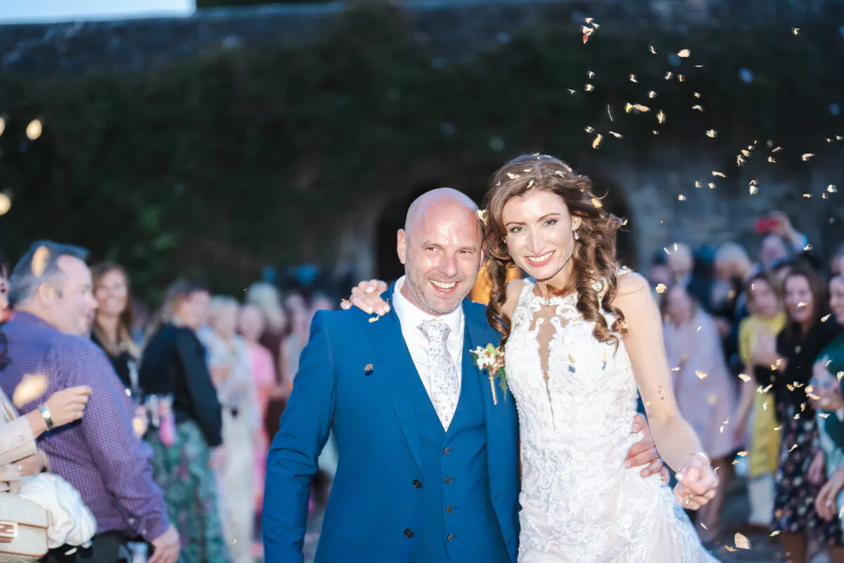 A joyful newlywed couple smiling amidst a shower of confetti as cheering guests celebrate around them outdoors.