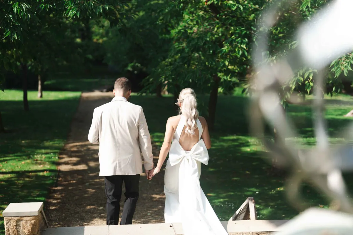 A newlywed couple walks hand in hand away from the camera down a sunny garden path lined with lush green trees.