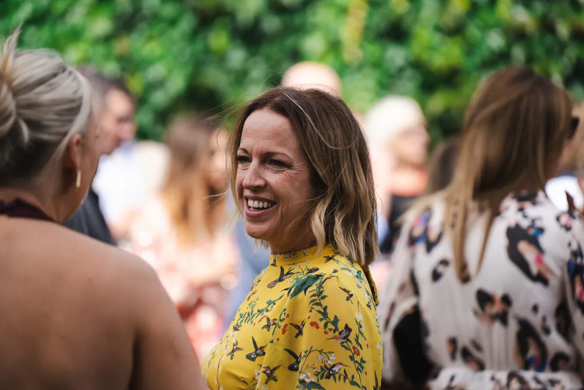 A smiling female wedding guest wearing a yellow floral print dress mingles and chats with other guests outdoors.