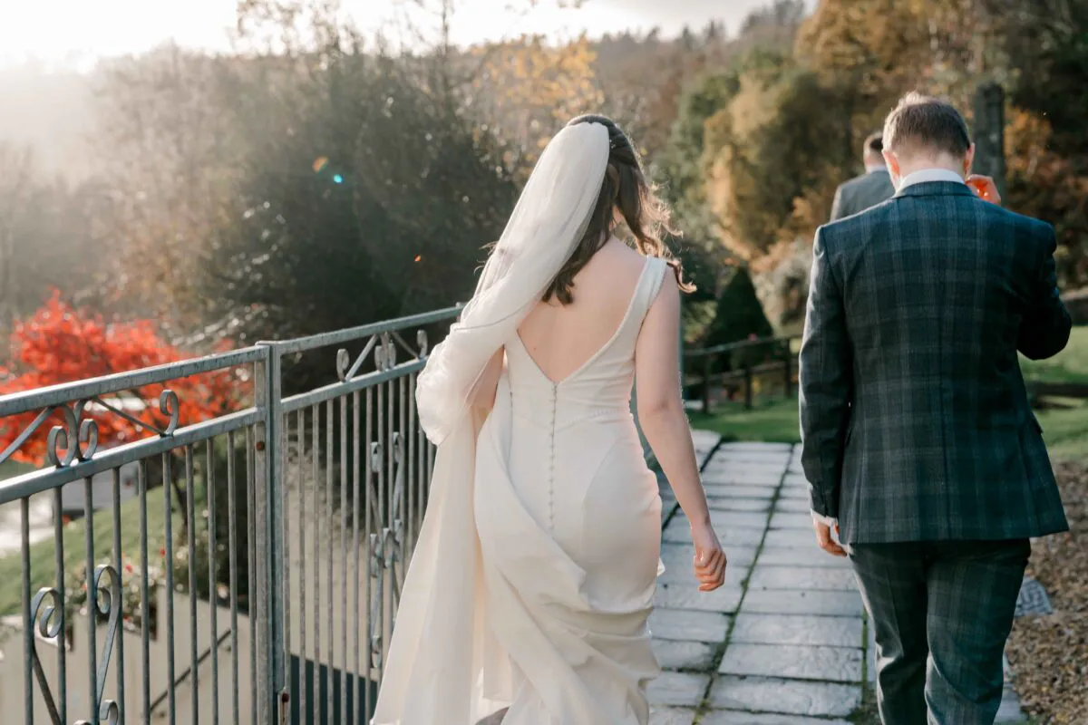 A bride in a sleek crepe gown with an open back and a long veil walks alongside the groom in a checked suit along an outdoor path during golden hour.