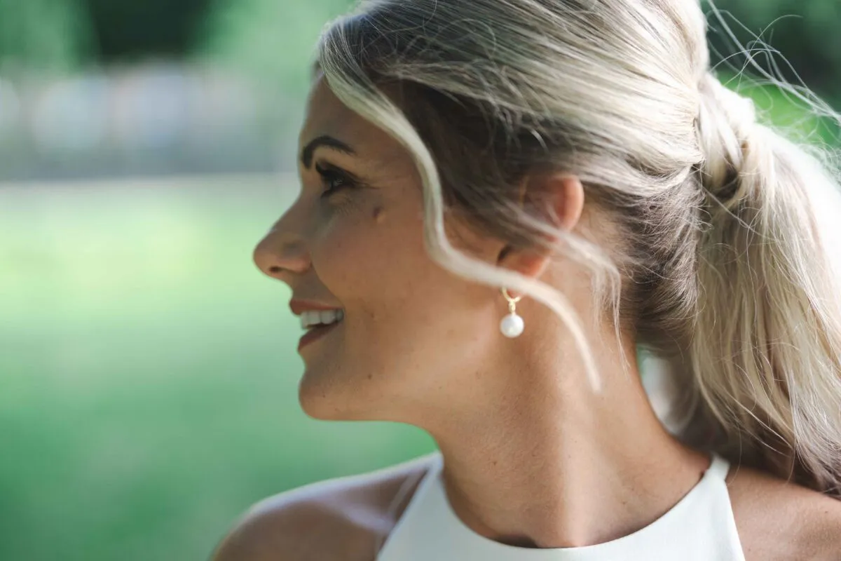 A close-up side profile of a smiling bride wearing her blonde hair in a high ponytail with a loose face-framing tendril and delicate pearl drop earrings.
