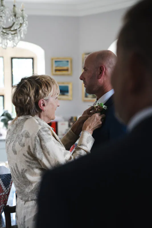 A mother gently pins a buttonhole onto her son's lapel as he prepares for his wedding day in a light-filled room.