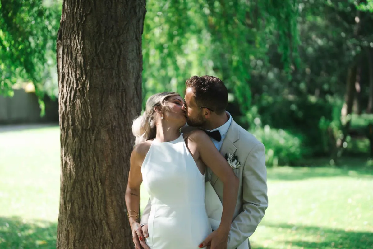 A bride in a sleek halter-neck gown and a groom in a beige suit share a tender kiss beneath a large tree in a sunlit woodland setting.