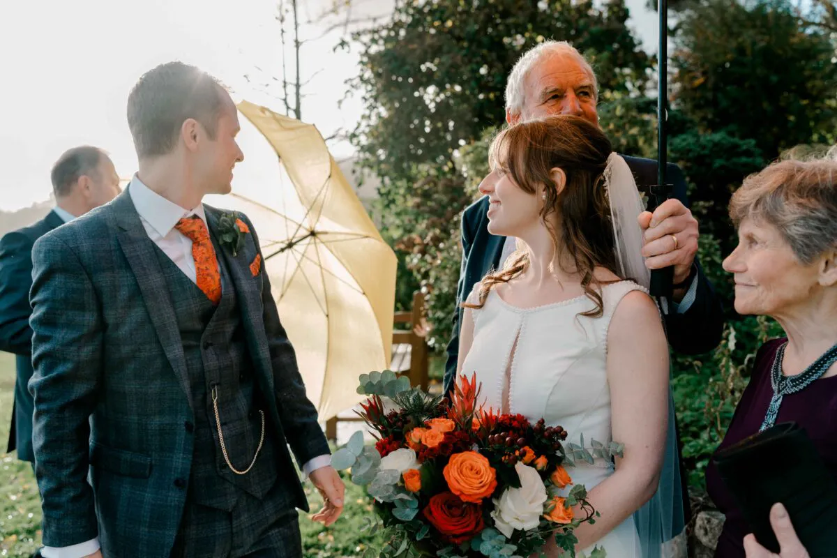 A radiant bride and groom exchange a tender glance outdoors during an autumn wedding, surrounded by family members holding yellow and clear umbrellas while backlit by warm afternoon sun.