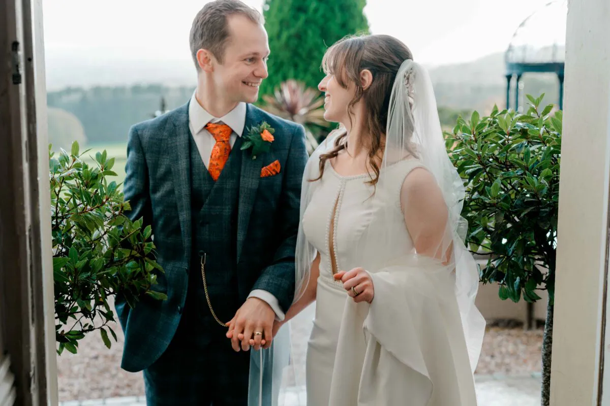 A smiling bride and groom hold hands and gaze at each other while framed by a doorway, with the groom wearing a tweed suit and the bride in a sleek satin gown and veil.